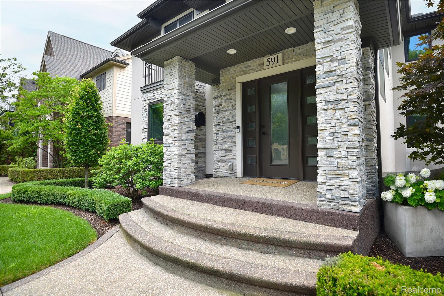 This image showcases the entryway of a modern home, featuring stone-clad pillars, a dark-framed front door with sidelights, and a multi-tiered concrete staircase leading up to the entrance. The landscaping is well-maintained with manicured hedges and green lawn, creating a welcoming and sophisticated curb appeal. The house number '591' is prominently displayed above the door.