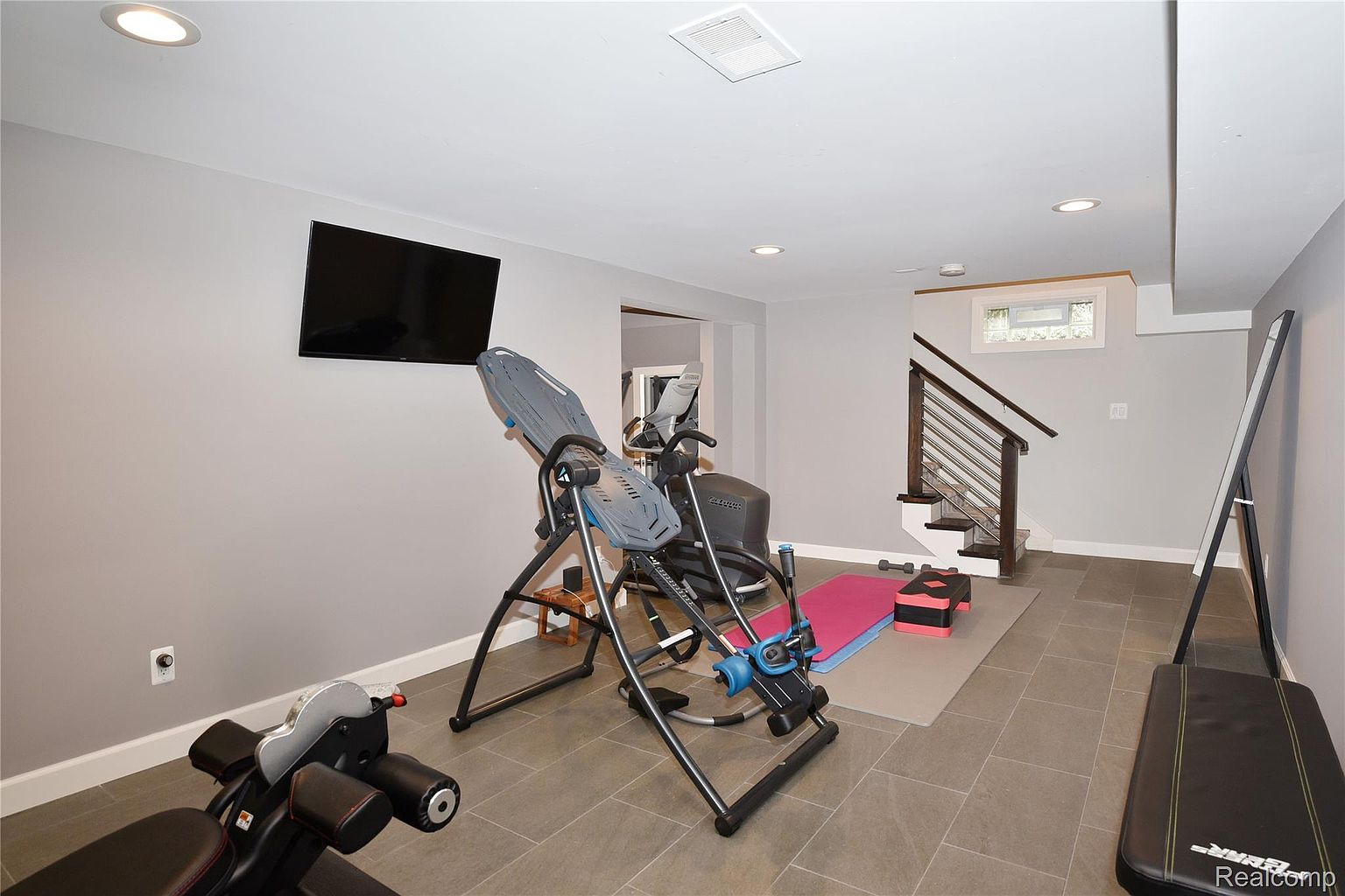 This is an interior shot of a home gym featuring various exercise equipment, including an inversion table, weight bench, and exercise bike. The room has tiled flooring, gray walls, and recessed lighting, creating a functional and well-lit workout space. A television is mounted on the wall, and stairs lead to another level.