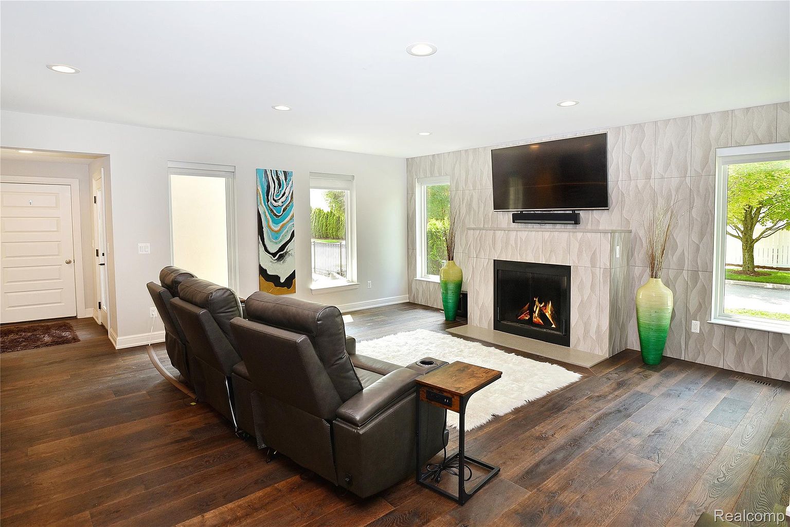 This is an interior shot of a living room featuring dark hardwood floors and a modern fireplace with a large television mounted above it. A comfortable-looking leather recliner sofa sits on a white shag rug, and natural light streams in through the windows, creating a warm and inviting atmosphere. The room is decorated with contemporary art and vases, adding to the stylish and comfortable ambiance.