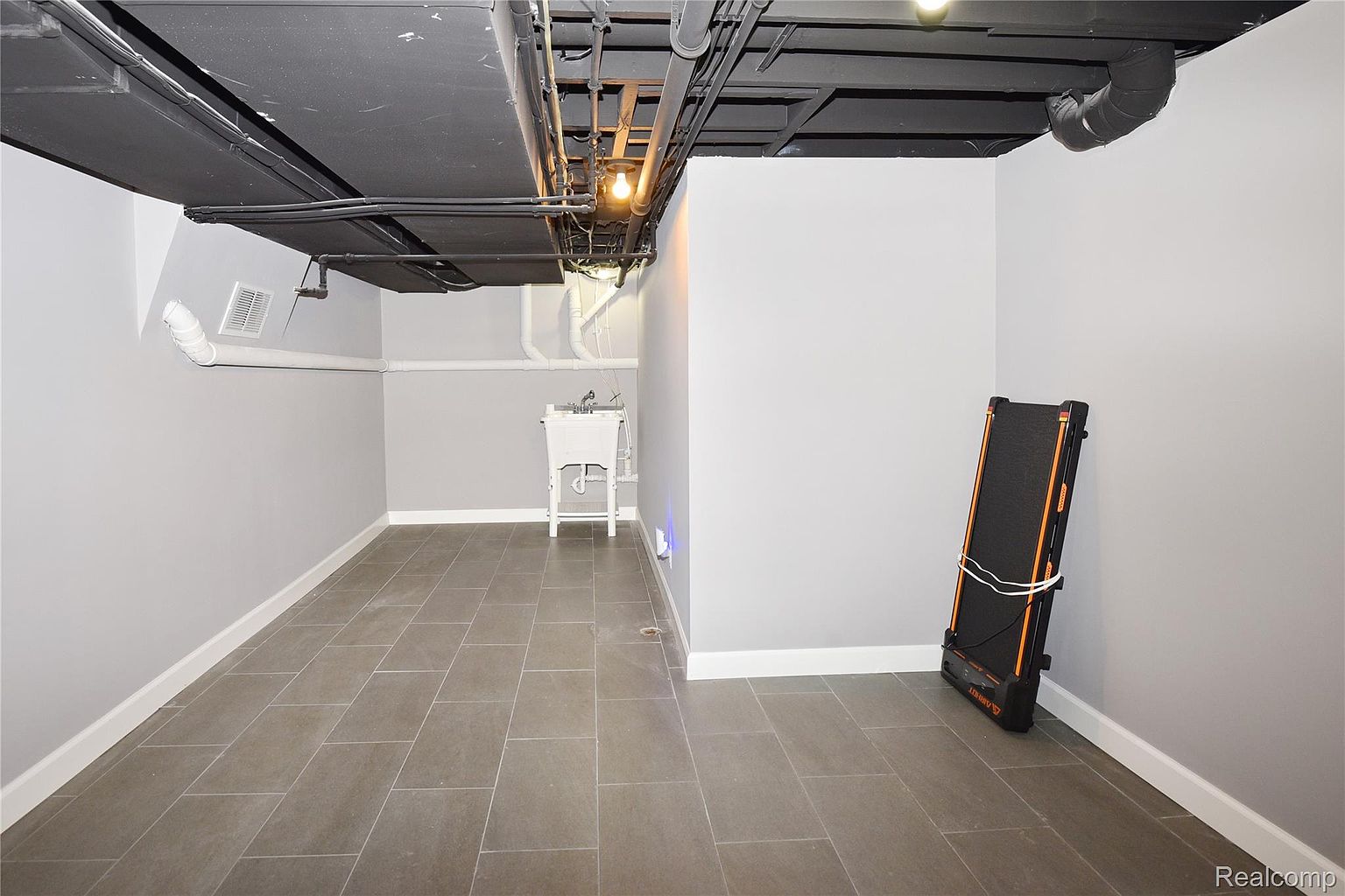 This is a finished basement featuring gray walls, a dark-painted ceiling with exposed pipes, and light gray tile flooring. A utility sink is visible in the corner, and a folded treadmill stands against the wall. The space appears clean and well-maintained, offering potential for various uses.