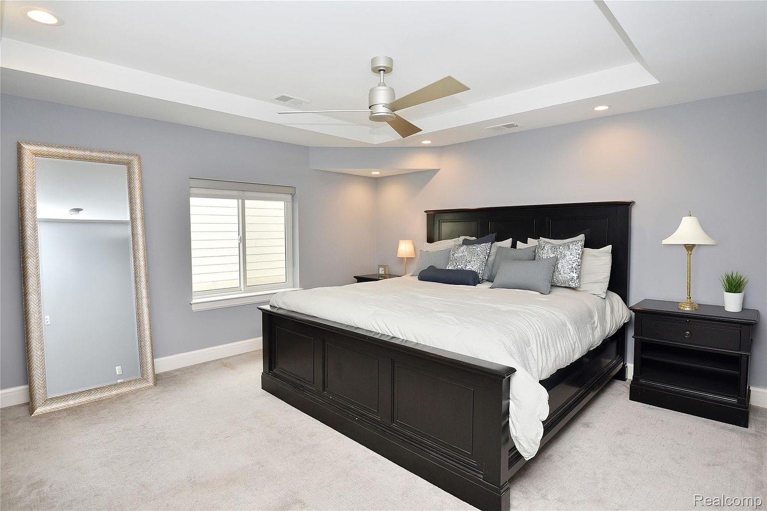 This is a primary bedroom featuring a large, dark wood bed with neutral bedding and decorative pillows. The room has a tray ceiling with recessed lighting and a ceiling fan. A full-length mirror stands against one wall, and a dark wood nightstand with a lamp sits beside the bed, creating a serene and inviting atmosphere.