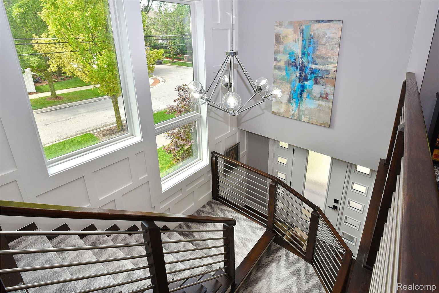 This is an interior shot of a modern home's hallway and staircase, viewed from above. The staircase features dark wood railings with horizontal metal bars and gray patterned flooring. A contemporary chandelier hangs above, and a large abstract painting adorns the wall. Large windows provide natural light, and the overall impression is one of modern elegance and spaciousness.