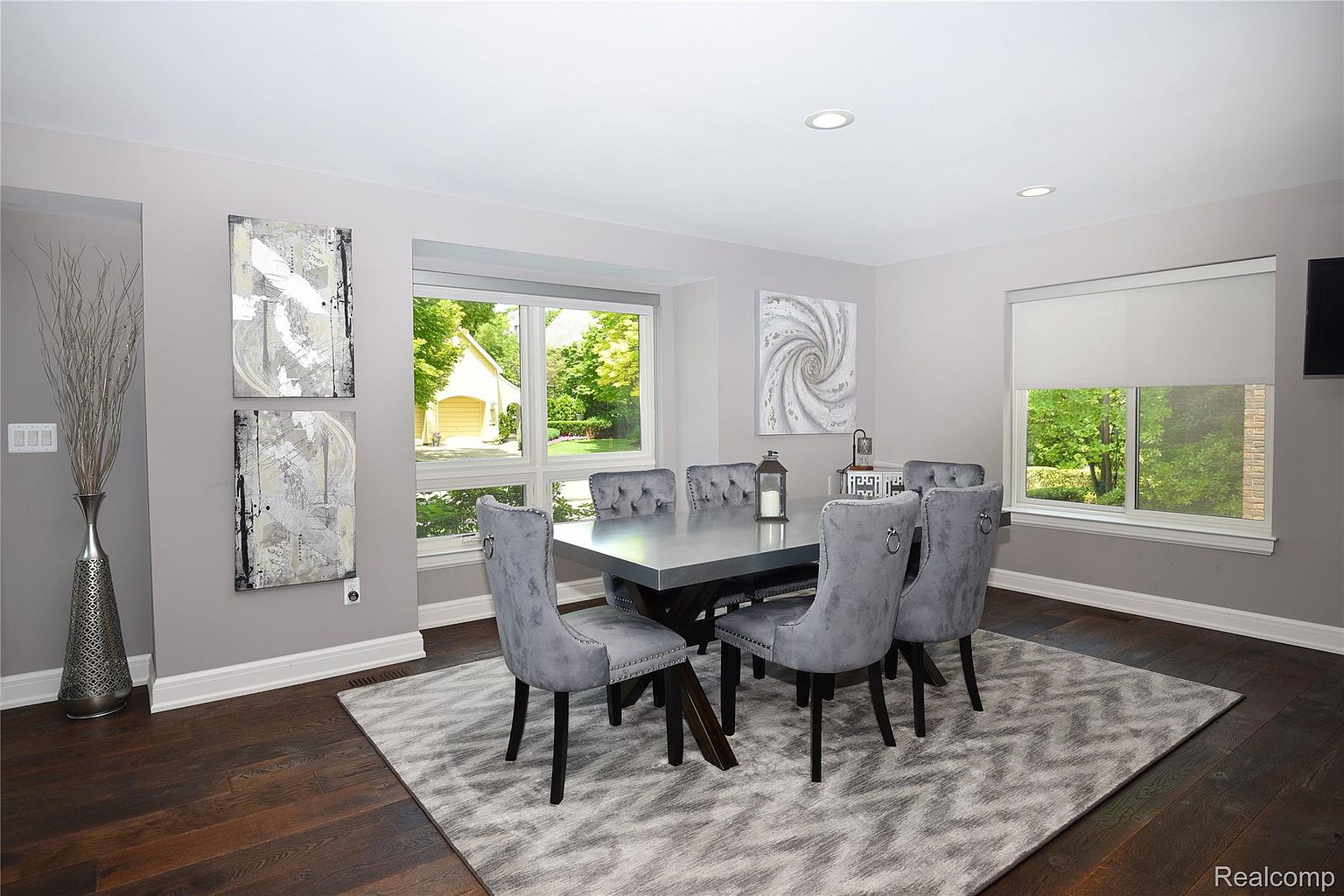 This is a dining room featuring a modern aesthetic with gray walls and dark hardwood floors. A rectangular dining table with a gray surface is surrounded by six gray upholstered chairs with button-tufted backs. The room is well-lit by natural light from a large window, complemented by recessed lighting, and a gray patterned rug anchors the space.