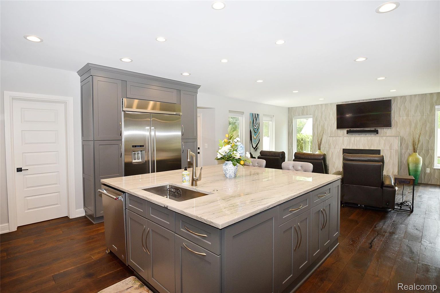 This is a well-lit, modern kitchen that seamlessly flows into the living area. The kitchen features a large island with a marble countertop, gray cabinetry, and stainless steel appliances, including a large refrigerator. The open floor plan enhances the sense of space and connectivity, making it ideal for entertaining.