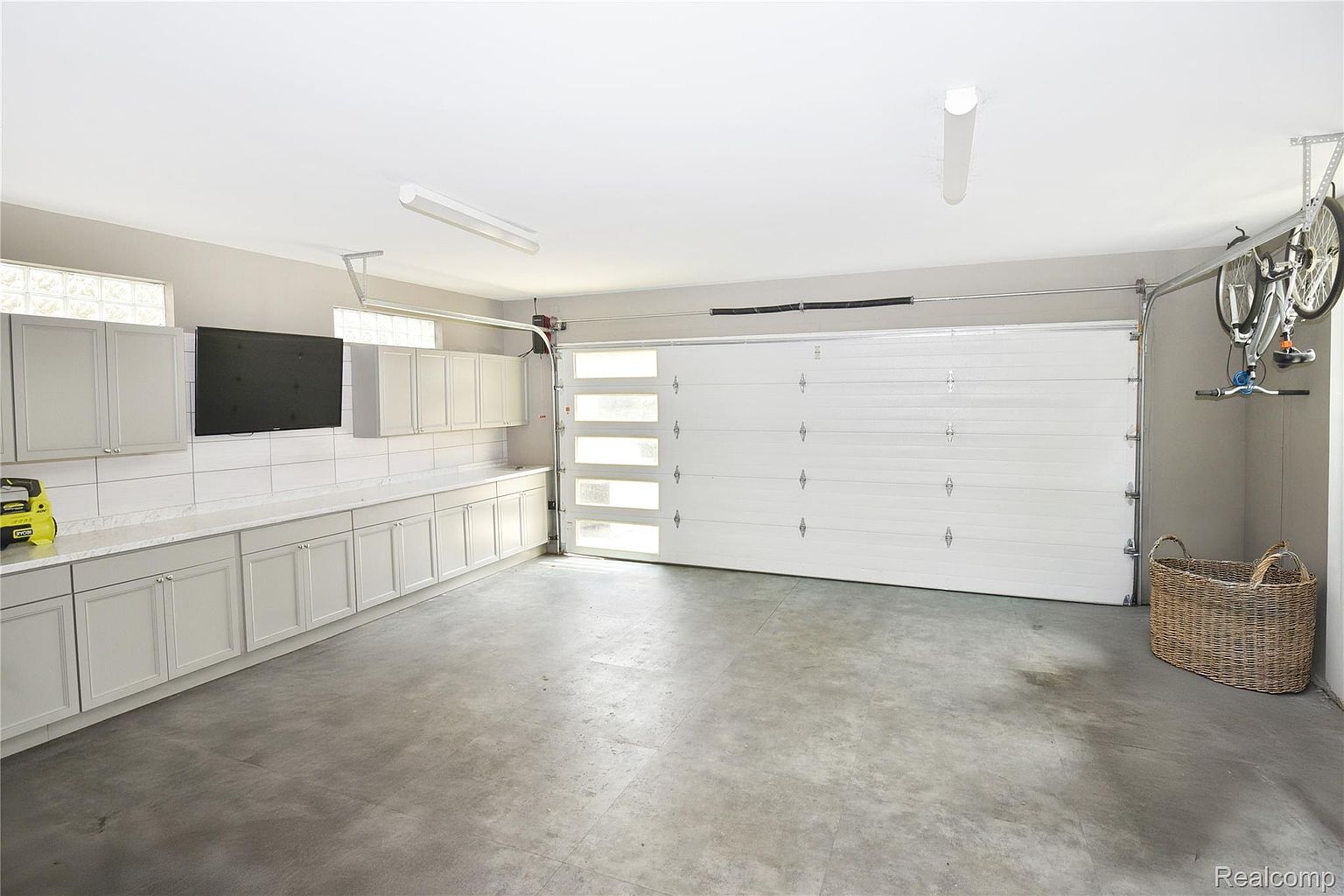 This is an interior shot of a well-organized garage. The garage features a white garage door with windows, light gray cabinets and countertops along one wall, and a gray concrete floor. A bicycle is mounted on the wall, and a wicker basket sits in the corner, adding a touch of warmth to the space.
