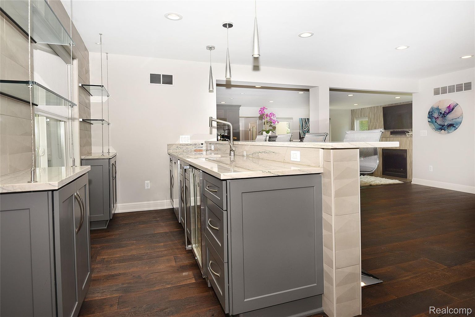 This interior shot showcases a modern kitchen area with dark wood flooring and gray cabinetry. A marble countertop extends into a bar area, illuminated by pendant lighting. The space opens into a living area, creating an open-concept feel, and the overall impression is stylish and inviting.