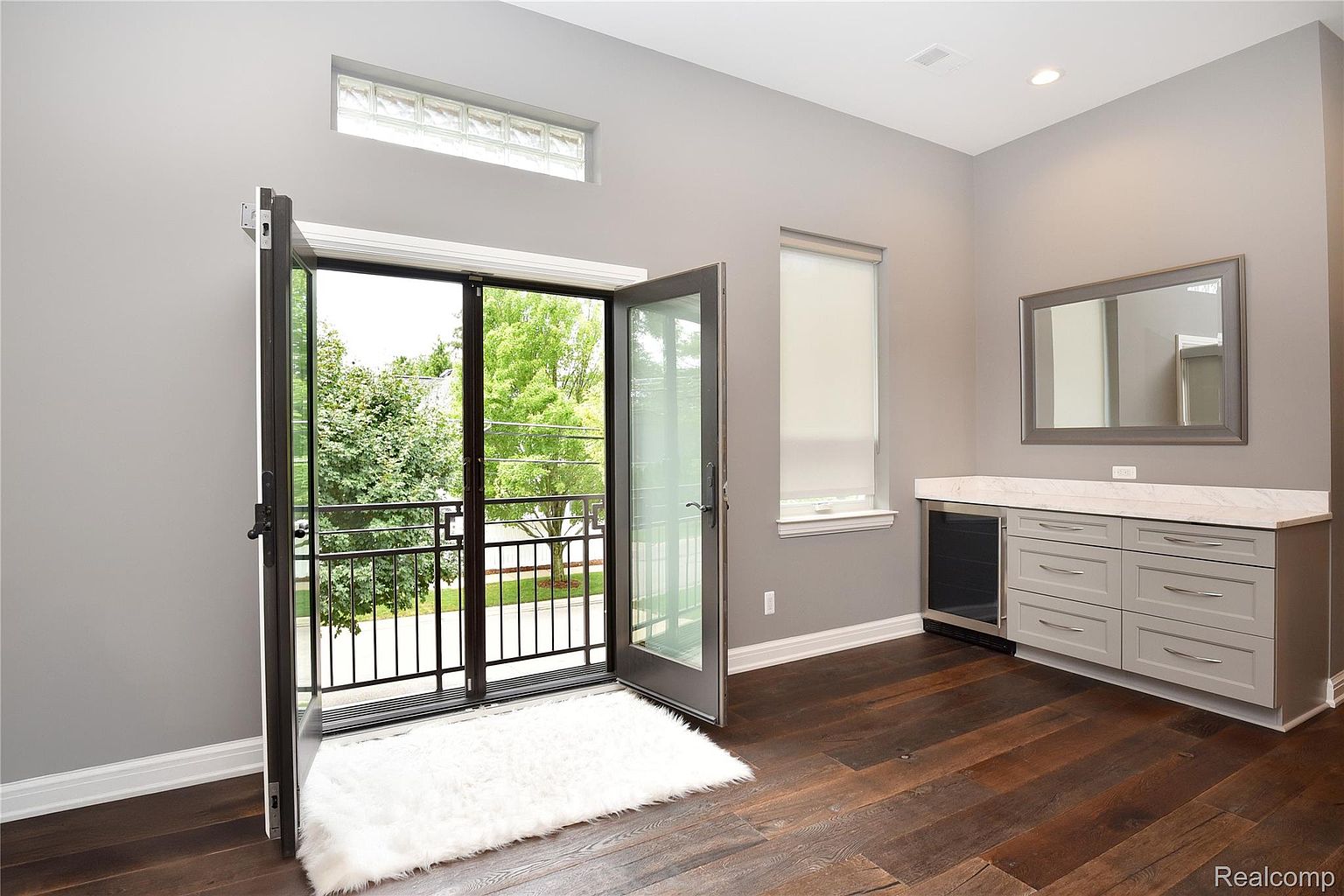 This primary bedroom features a modern design with gray walls and dark hardwood floors. A balcony is accessible through glass doors, providing natural light and a view of the outdoors. The room also includes a built-in bar area with a marble countertop and a mini-fridge, adding a touch of luxury and convenience.