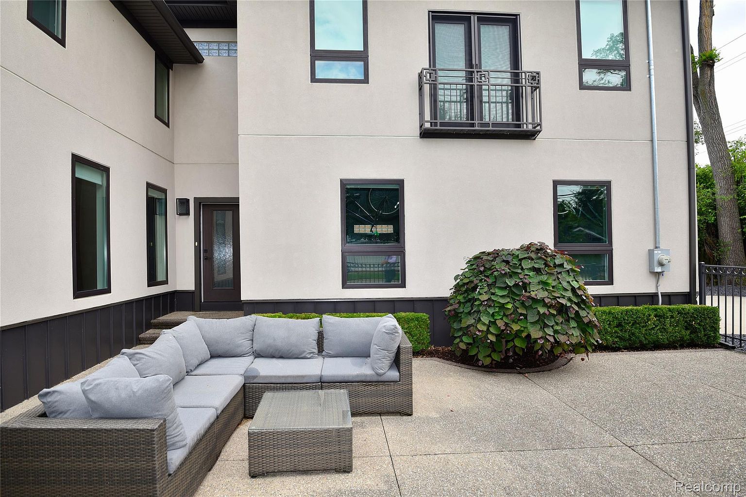 This image showcases a modern home's patio area, featuring a stylish gray wicker sectional sofa and coffee table set on a concrete patio. The exterior of the house is a light stucco with dark-framed windows and a small balcony above. Lush greenery surrounds the patio, adding a touch of nature to the contemporary design.