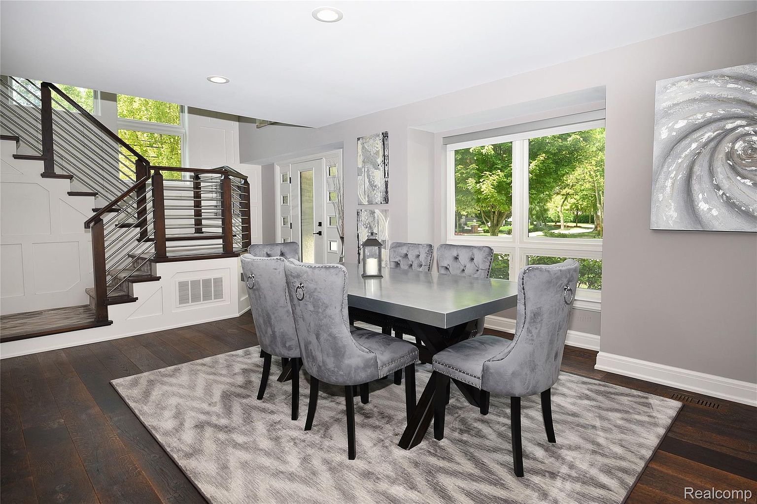 This is an interior shot of a dining room featuring a modern aesthetic. The room is furnished with a gray dining table and six matching velvet chairs. A large window provides natural light, and a contemporary artwork hangs on the wall, complementing the room's color scheme. The flooring is dark wood, and a patterned rug anchors the dining set, adding texture to the space.