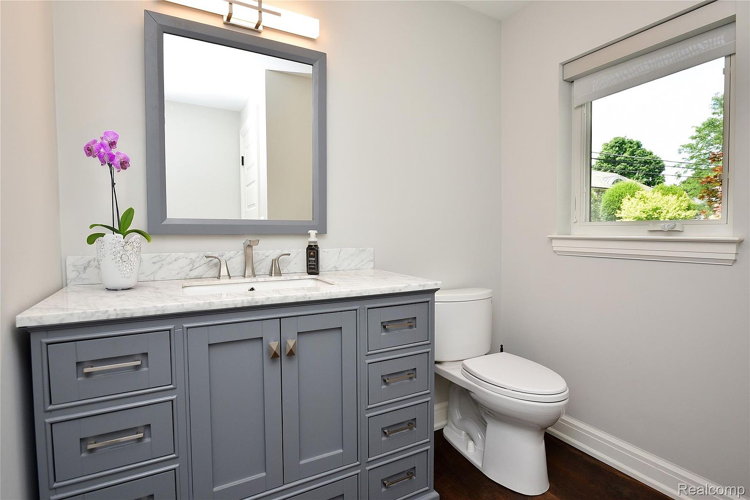 This is a well-lit guest bathroom featuring a gray vanity with a marble countertop, a modern sink and faucet, and a framed mirror with a light fixture above. A white toilet sits adjacent to the vanity, and a window provides natural light. The overall impression is clean, stylish, and functional.