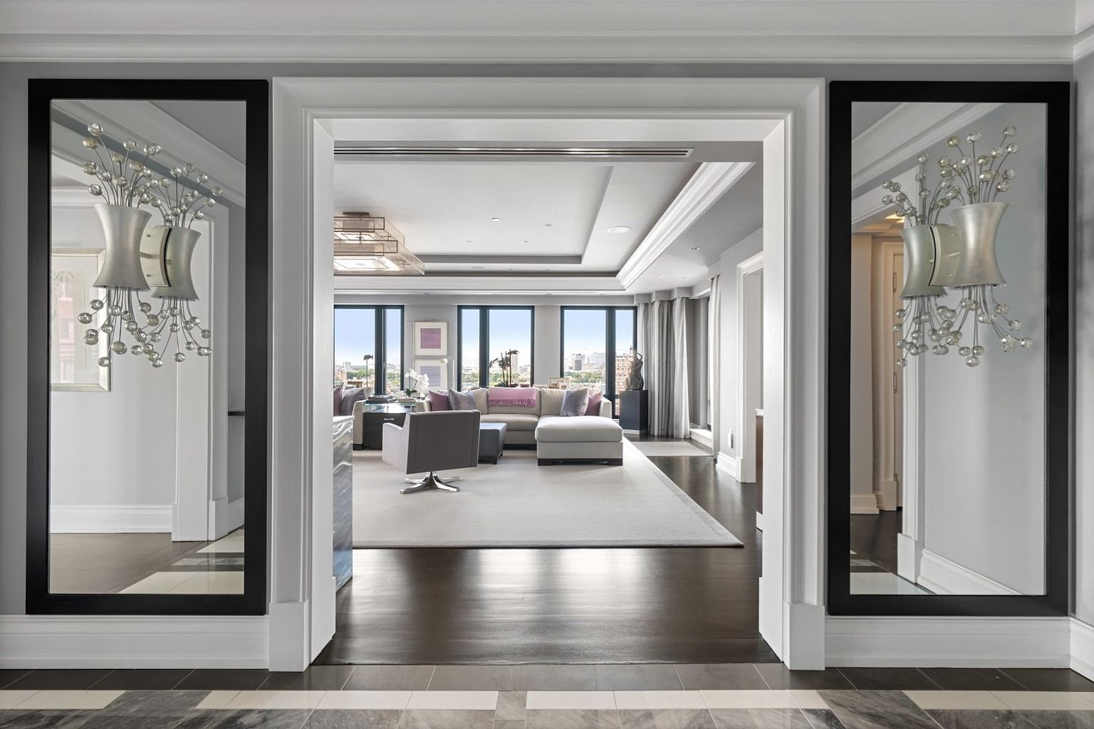 This image showcases a luxurious living room viewed through an elegant doorway, featuring a neutral color palette with pops of purple and gray. The room is furnished with a large sectional sofa, a modern armchair, and a plush area rug, while large windows offer city views. Decorative mirrors with sconces flank the doorway, adding to the room's sophisticated ambiance.