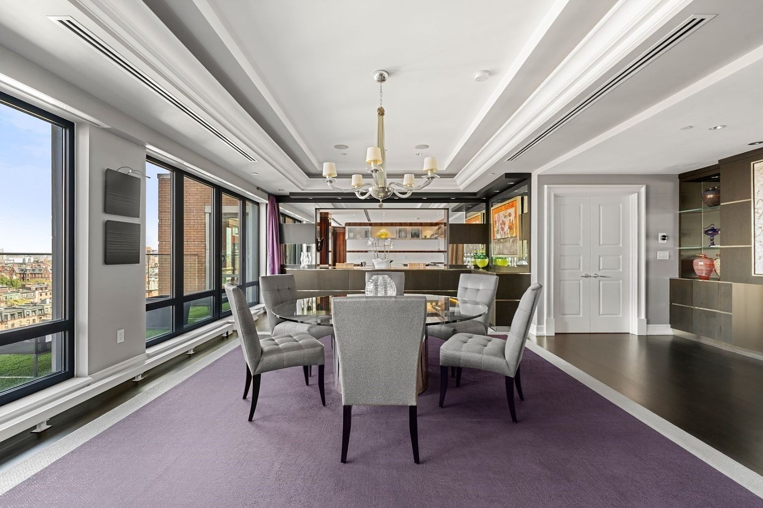 This interior shot showcases a sophisticated dining room with a round glass table surrounded by four upholstered chairs on a purple rug. A modern chandelier hangs above the table, and large windows offer a city view, enhancing the room's elegance. The room is well-lit and appears spacious, perfect for entertaining.