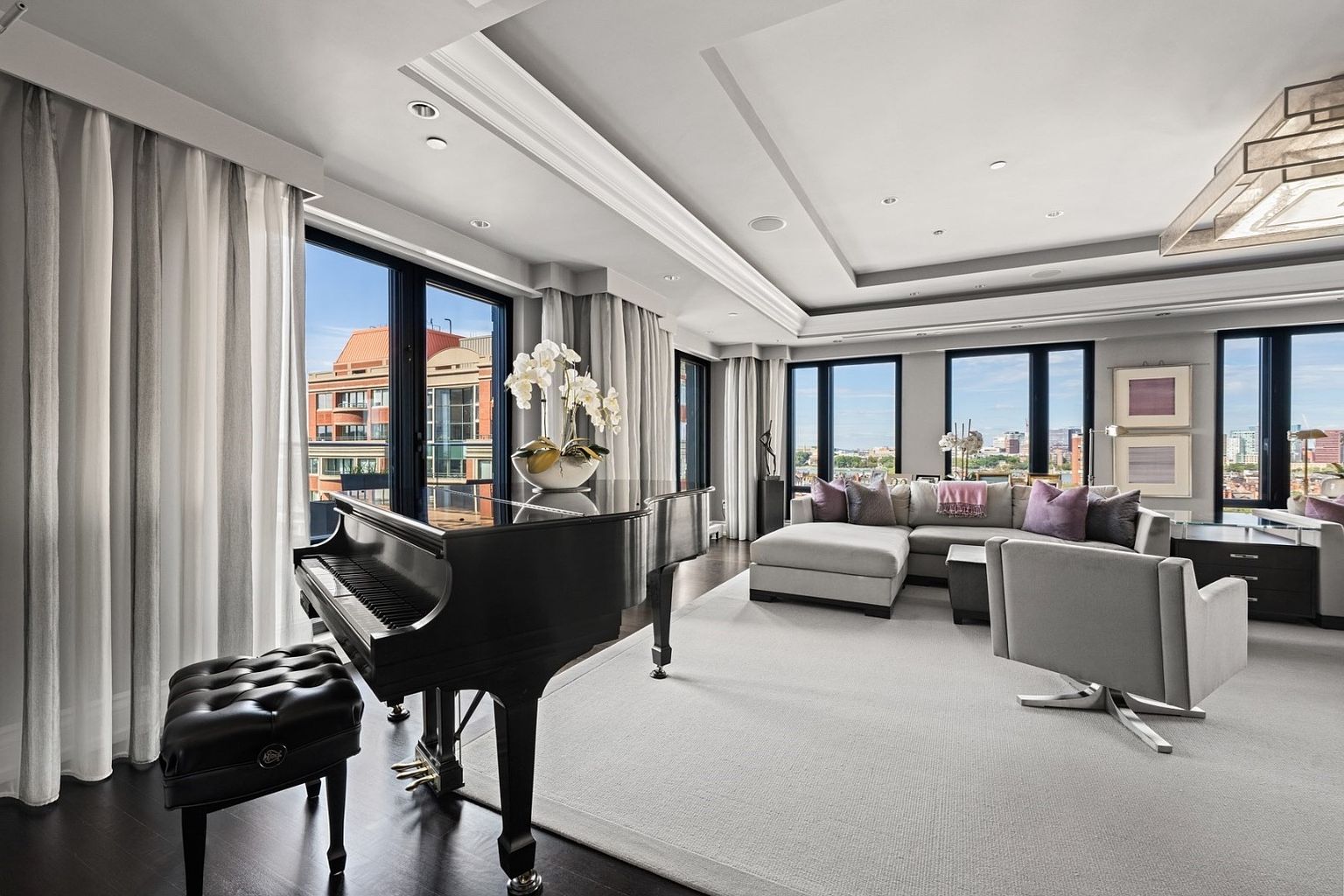 This is a luxurious living room featuring a grand piano, a plush sectional sofa with decorative pillows, and a stylish gray armchair. Large windows offer city views, complemented by elegant curtains and a sophisticated lighting fixture. The room exudes a sense of opulence and comfort, perfect for upscale living.
