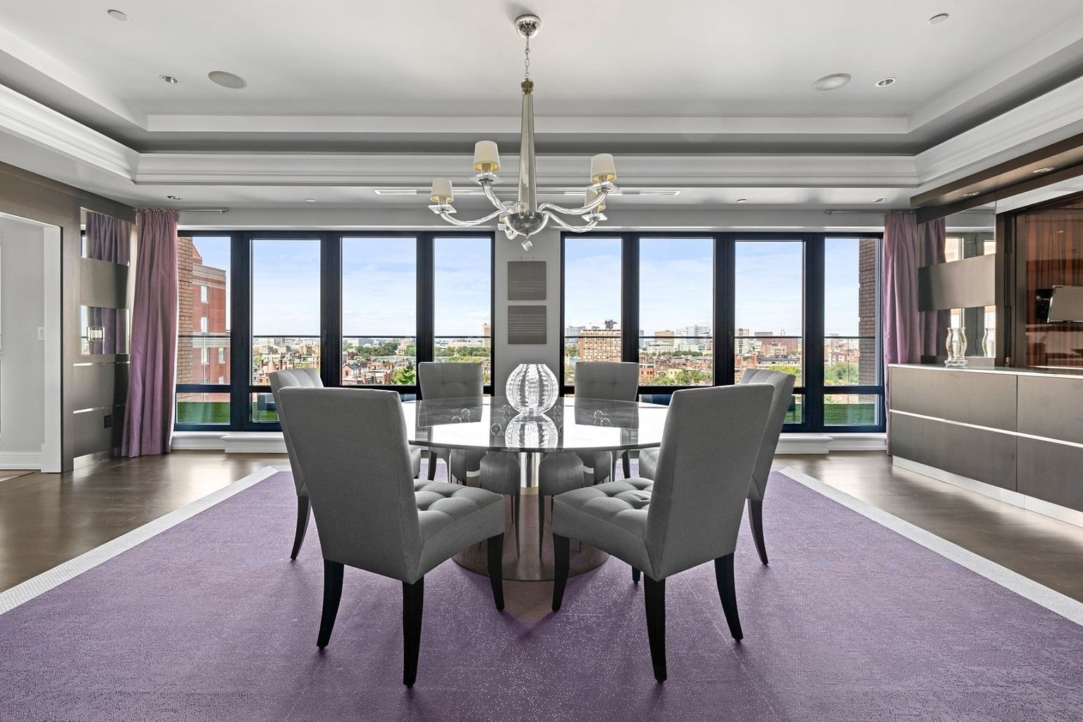 This is a dining room featuring a round glass table with six gray upholstered chairs. A modern chandelier hangs above the table, and large windows offer a city view. A purple rug defines the dining area, and the overall style is elegant and contemporary.