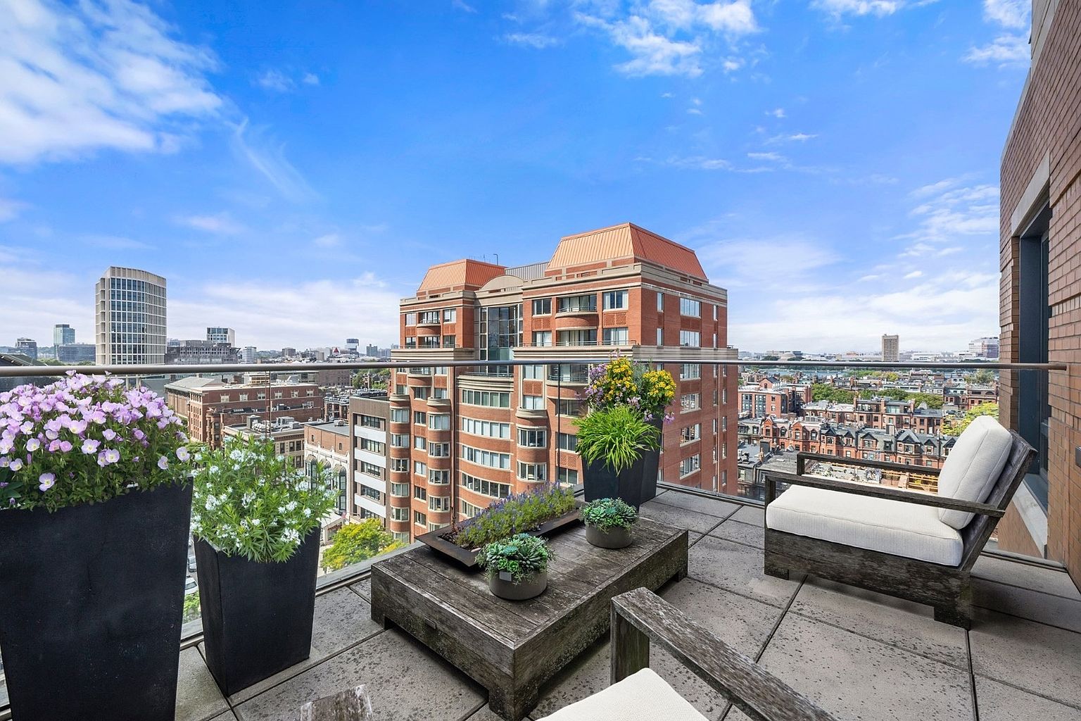 This image showcases a luxurious balcony with a stunning cityscape view. The outdoor space is furnished with comfortable seating, including chairs with white cushions, and a rustic wooden coffee table adorned with potted plants. The balcony is lined with a modern glass railing, providing an unobstructed view of the surrounding buildings and sky, creating an inviting and upscale outdoor living area.