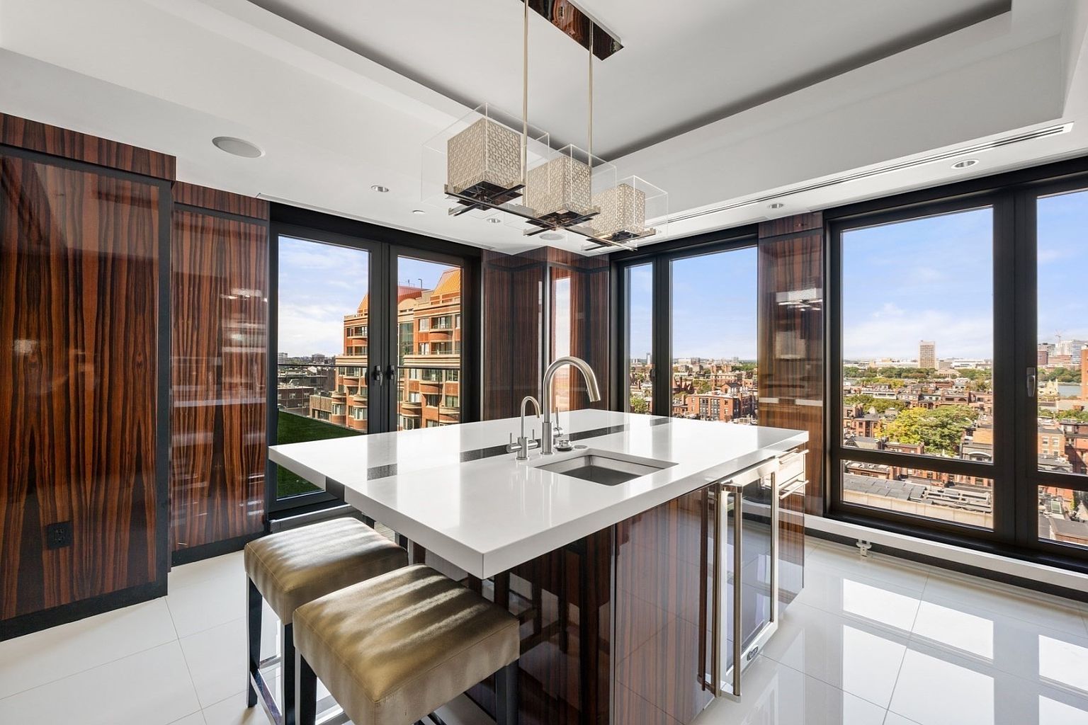 This is a modern kitchen with sleek, dark wood cabinetry and a large white countertop island featuring a sink and seating. The room is well-lit with contemporary pendant lighting and natural light streaming through large windows that offer a city view. The glossy white tile floor adds to the clean and luxurious feel of the space.
