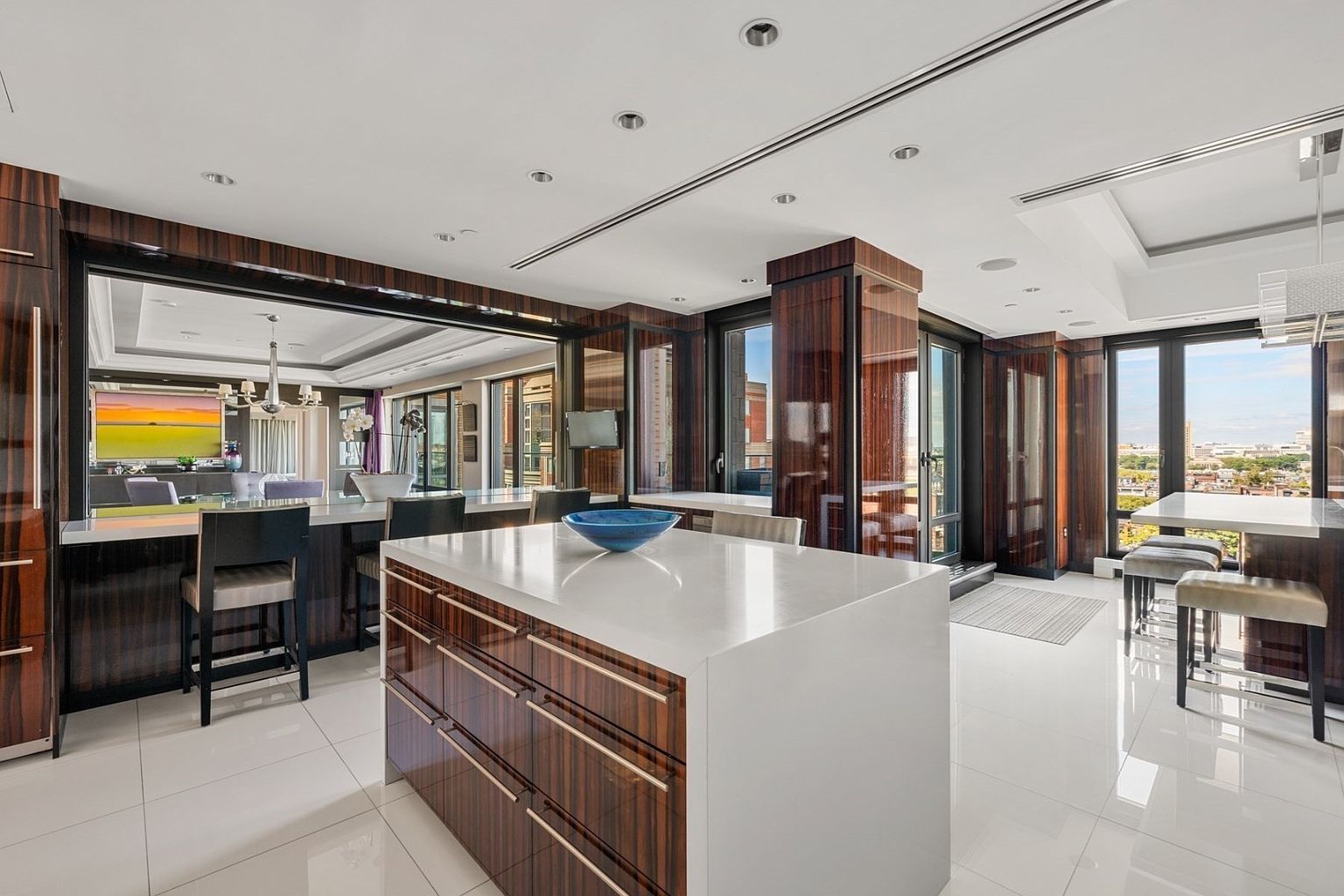 This is a modern, high-end kitchen featuring sleek, dark wood cabinetry with metallic accents and a large white countertop island. The kitchen boasts an open layout with a view into the living area through a large opening, and floor-to-ceiling windows provide ample natural light and city views. The glossy white tile flooring adds to the contemporary aesthetic.