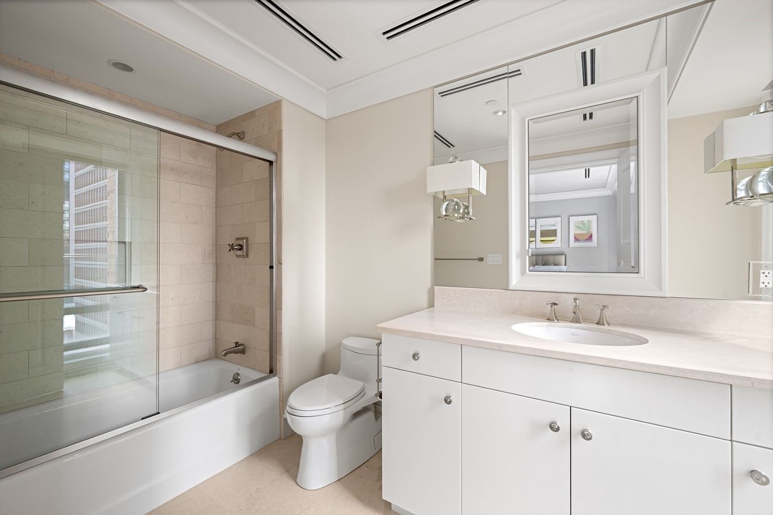 This is a well-lit bathroom featuring a bathtub with a glass shower door, a white toilet, and a vanity with white cabinets and a light-colored countertop. A large mirror hangs above the vanity, reflecting the room and enhancing the sense of space. The overall impression is clean, modern, and functional.