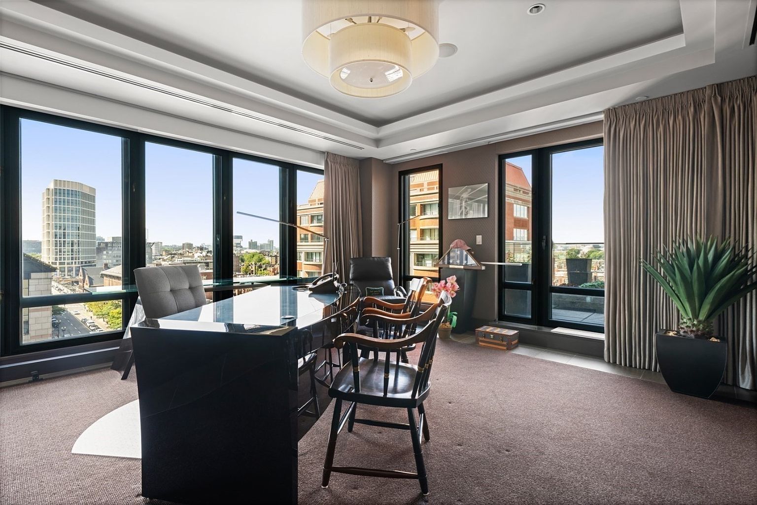 This is an interior shot of a sophisticated office space featuring a large, glossy black desk as the focal point, complemented by a gray office chair and several wooden guest chairs. The room is well-lit by natural light streaming through large windows that offer city views, and the space is decorated with a modern chandelier and a potted plant, creating a professional yet inviting atmosphere.