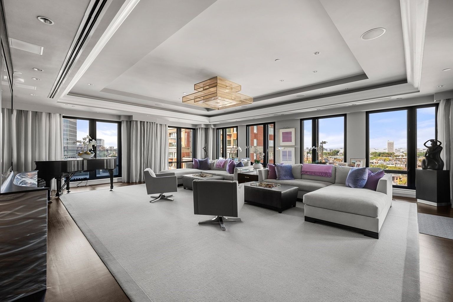 This is a spacious and luxurious living room featuring a large sectional sofa with purple accents, two modern swivel chairs, and a grand piano. The room is well-lit with natural light from large windows offering city views, complemented by a stylish chandelier. The overall impression is one of elegance and sophistication, perfect for upscale living.