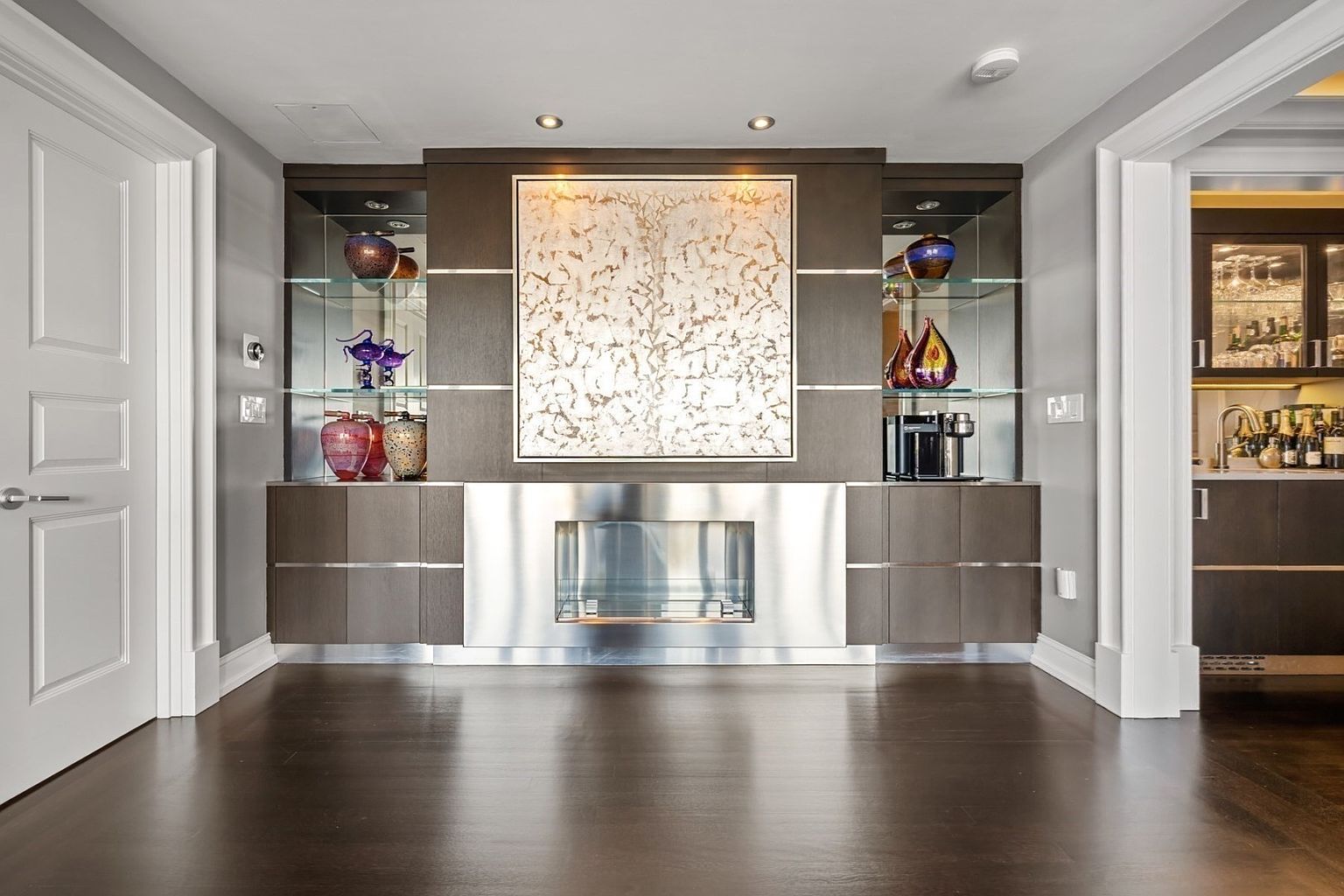 This interior shot showcases a modern living room with a built-in entertainment center featuring glass shelves, artwork, and a sleek fireplace. The dark hardwood floors provide a rich contrast to the lighter walls and cabinetry. The room exudes a sophisticated and contemporary ambiance, perfect for entertaining or relaxing.