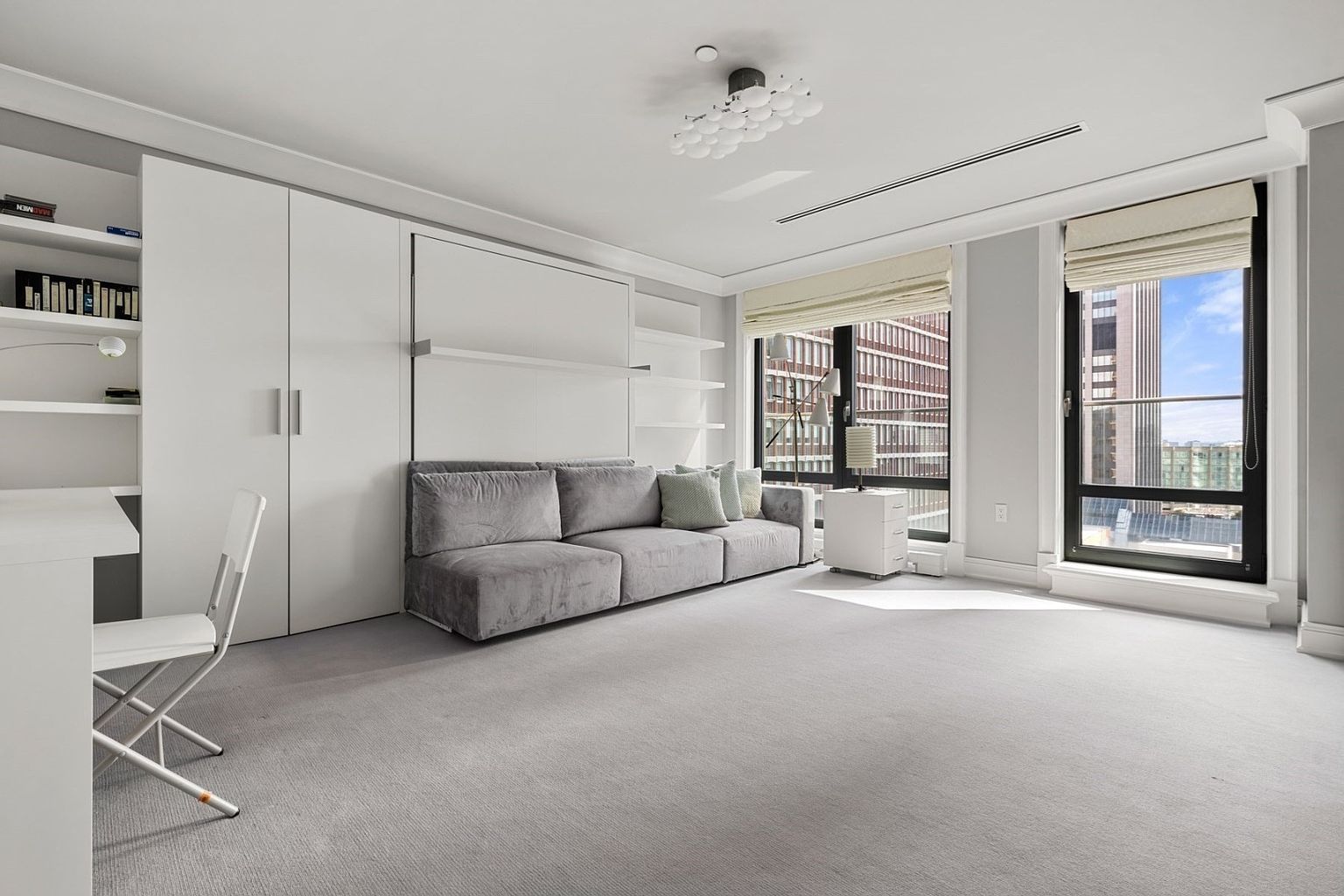 This is a bright and airy guest bedroom featuring a gray sofa bed, built-in shelving, and a desk area. Large windows offer city views, and the neutral color palette creates a calming atmosphere. The room is well-lit and appears spacious, making it an inviting space for guests.