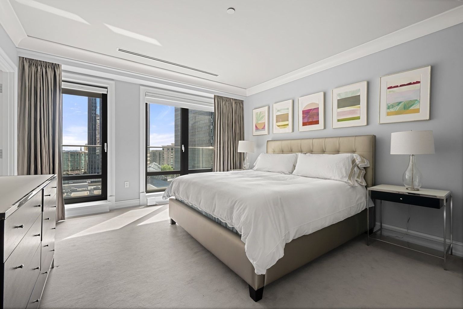 This is a well-lit primary bedroom featuring a large bed with white linens, flanked by a nightstand and a dresser. The room has large windows offering city views, complemented by neutral-toned curtains and artwork above the bed. The overall impression is one of modern elegance and comfort.