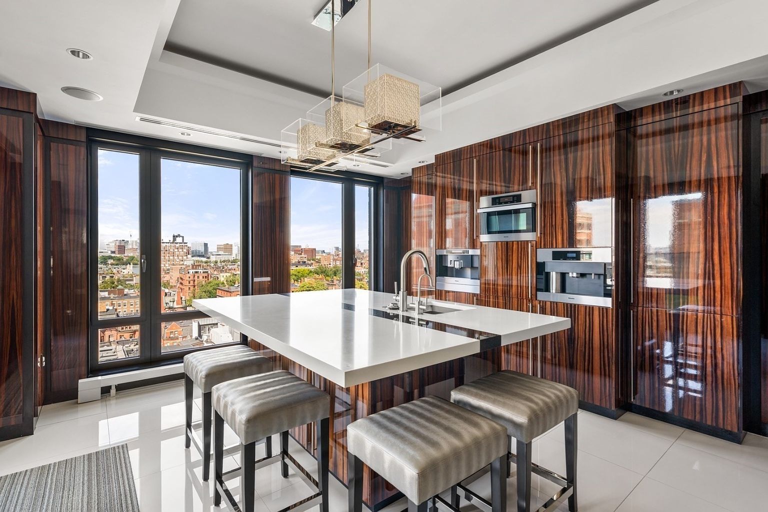 This is a modern kitchen featuring dark wood cabinetry with integrated stainless steel appliances. A large white countertop island with seating is the focal point, illuminated by a contemporary chandelier. Large windows offer a city view, enhancing the luxurious feel of the space.