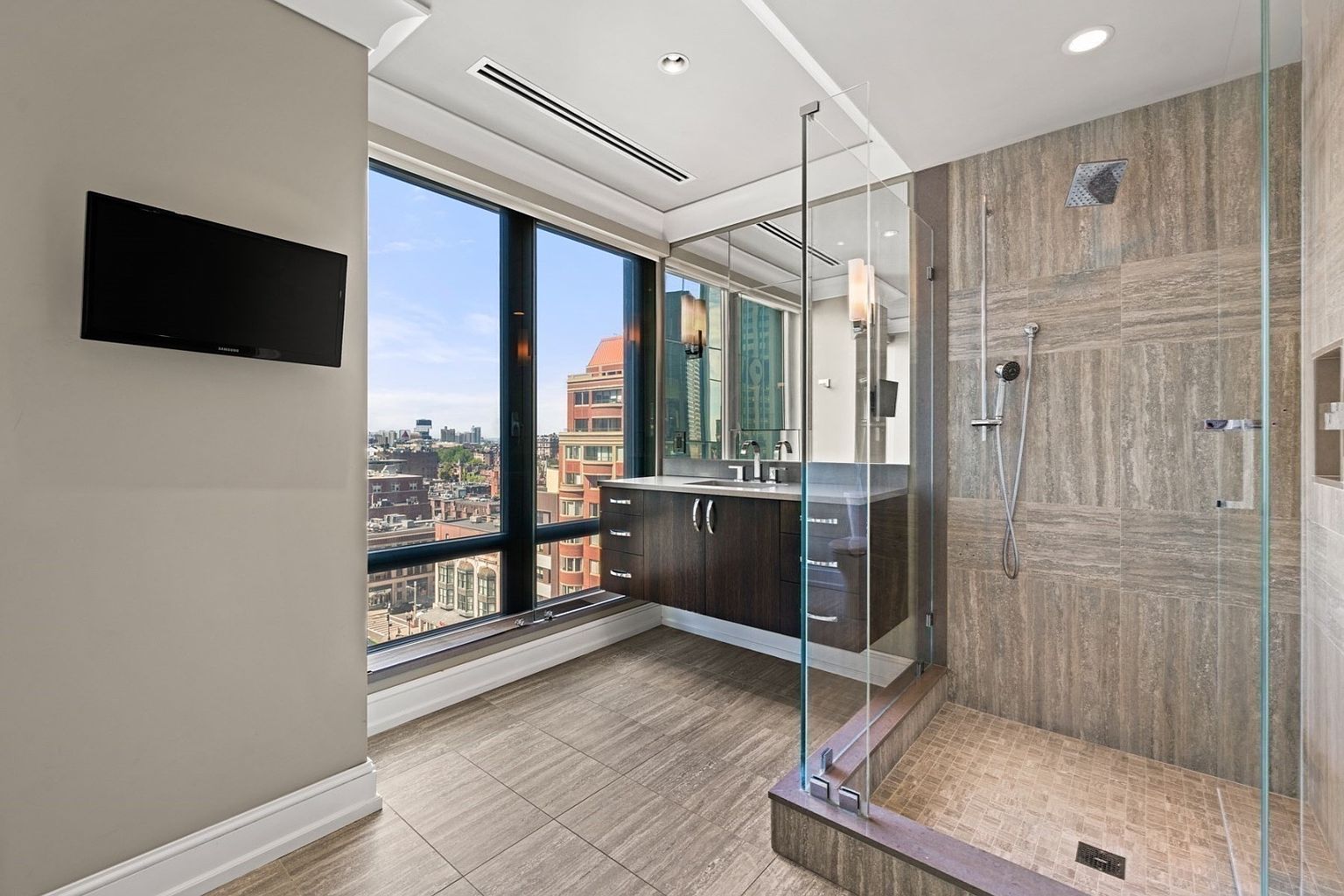 This is a luxurious primary bathroom featuring a modern design with a large window offering city views. The bathroom includes a sleek vanity with dark wood cabinetry, a glass-enclosed shower with tiled walls, and a wall-mounted television. The overall impression is one of sophistication and comfort.