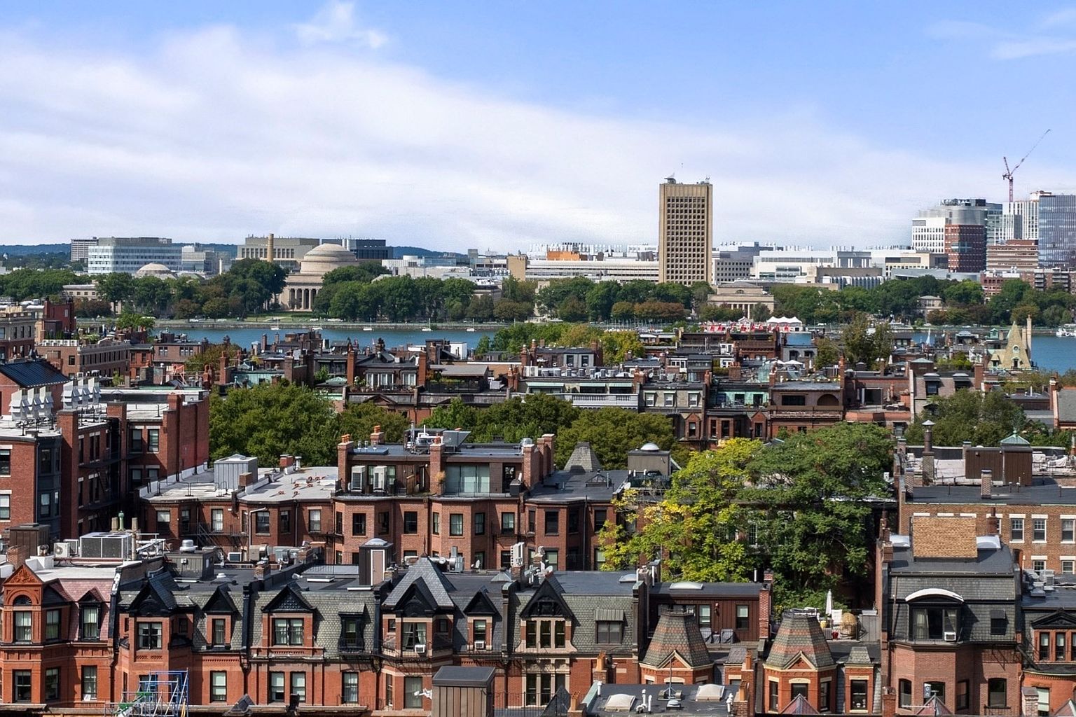 This aerial view showcases a dense urban landscape with rows of brick townhouses, interspersed with mature trees. In the background, a river separates the residential area from a skyline featuring various institutional and commercial buildings, including a prominent tower. The scene is bathed in natural light under a partly cloudy sky, creating a vibrant and appealing cityscape.