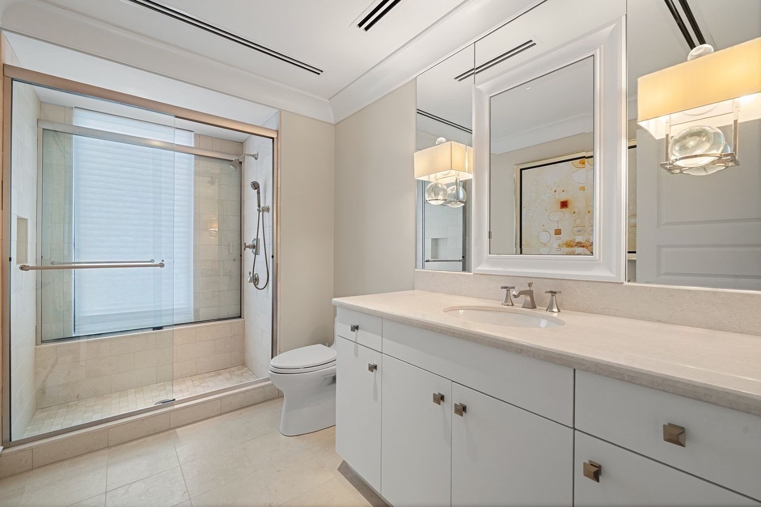 This is a well-lit primary bathroom featuring a glass-enclosed shower with tiled walls and a built-in shower niche. A white vanity with a light countertop and modern hardware is complemented by a large mirror and decorative lighting. The overall impression is clean, bright, and luxurious.