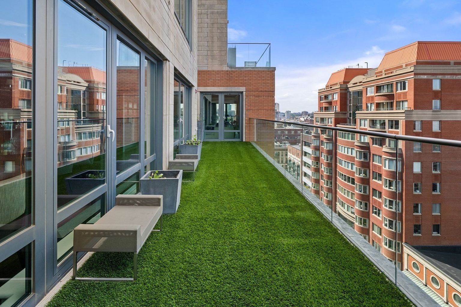 This image showcases a luxurious balcony with artificial turf, offering a unique outdoor space. The balcony features modern glass railings and a sleek bench, providing a comfortable seating area. The view includes neighboring buildings and a glimpse of the cityscape, adding to the urban appeal.