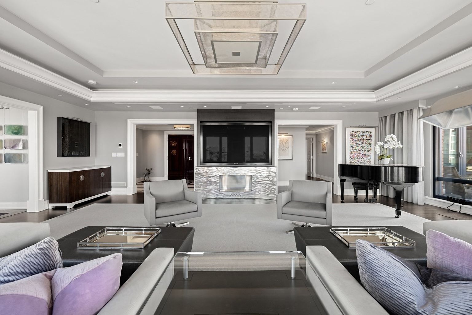 This is a luxurious living room featuring a modern design with a neutral color palette. The room includes a large television mounted above a decorative fireplace, two gray armchairs, and a grand piano. The space is well-lit with a unique ceiling light fixture and natural light from the windows, creating an elegant and inviting atmosphere.