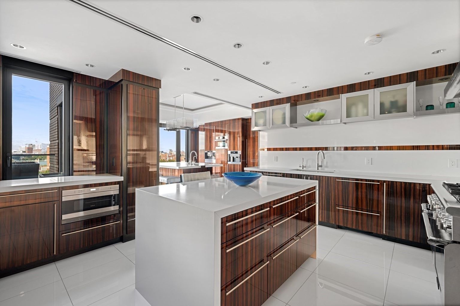 This is a modern kitchen featuring dark wood cabinetry with sleek, horizontal hardware and white countertops. A large island with a white countertop and dark wood base stands in the center, topped with a blue bowl. Stainless steel appliances are integrated into the cabinetry, and large windows offer a city view, creating a luxurious and functional space.