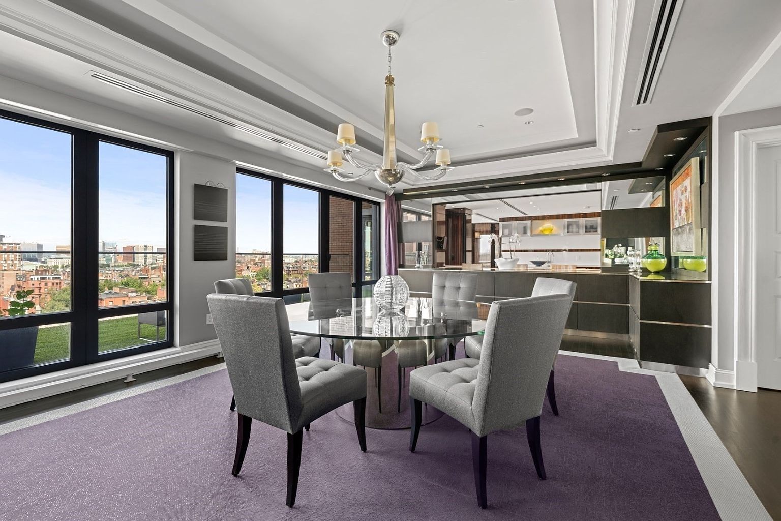 This is an interior shot of a dining room featuring a round glass table surrounded by six gray upholstered chairs. A modern chandelier hangs above the table, and large windows offer a city view. The room is decorated with a purple rug, and the overall style is elegant and contemporary.