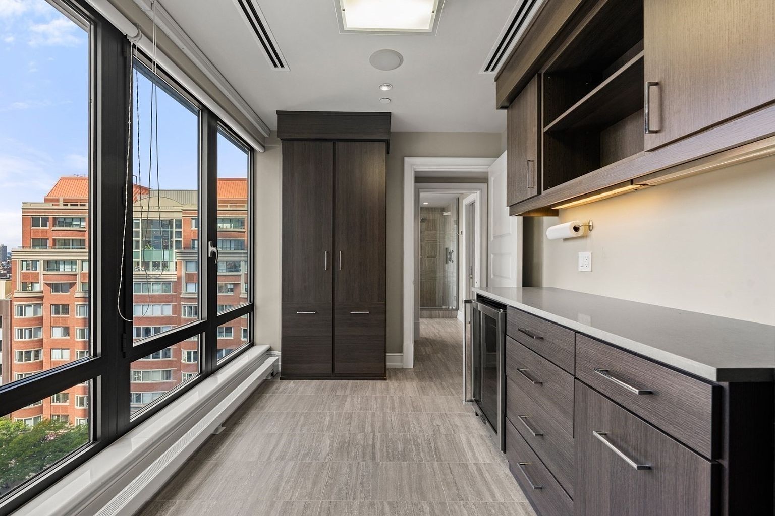 This interior shot showcases a modern hallway featuring dark wood cabinetry and drawers, complemented by light gray countertops and flooring. A large window provides ample natural light and a view of the city, while a doorway leads to another room, possibly a bathroom. The space is clean, well-lit, and appears to be part of a high-end apartment or condo.