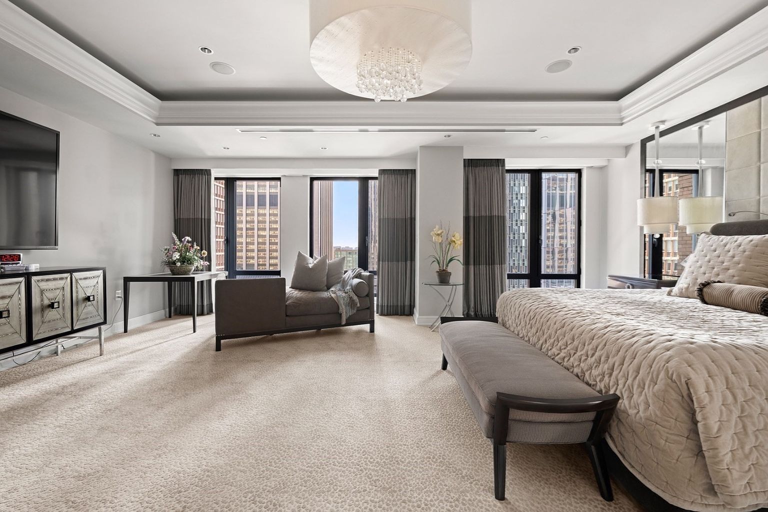 This is a luxurious primary bedroom featuring a large bed with a quilted cover, a seating area with a chaise lounge, and floor-to-ceiling windows offering city views. The room is decorated in neutral tones with gray accents, and a modern chandelier hangs from the ceiling. The overall impression is one of elegance and comfort.
