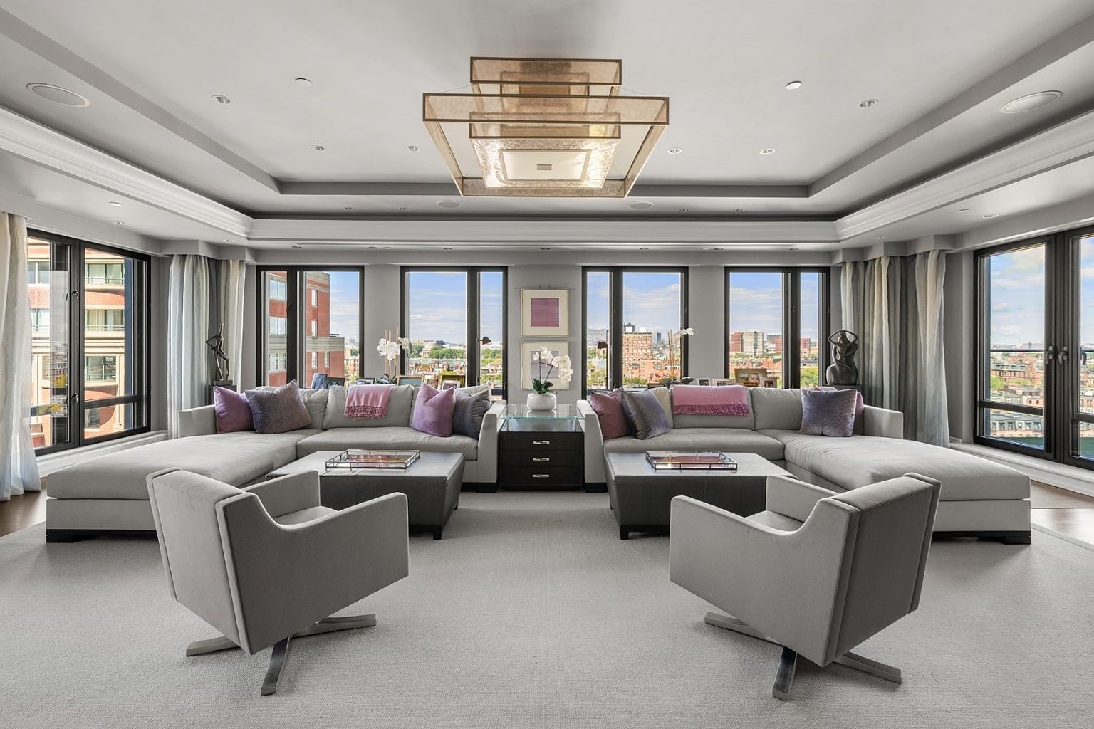 This is a spacious and luxurious living room featuring a large sectional sofa with decorative pillows, two modern armchairs, and a contemporary chandelier. The room is well-lit with natural light from the large windows, offering city views. The neutral color palette and elegant furnishings create a sophisticated and inviting atmosphere.