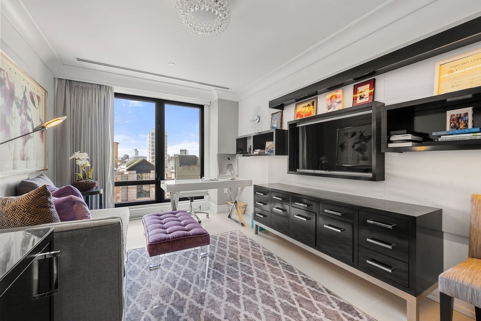 This is an interior shot of a modern office space featuring a white desk, a purple ottoman, and a dark wood cabinet with a mounted television. The room has a large window offering a city view, and the decor includes artwork, books, and decorative items on floating shelves. The overall impression is stylish and functional, suitable for a professional or creative workspace.