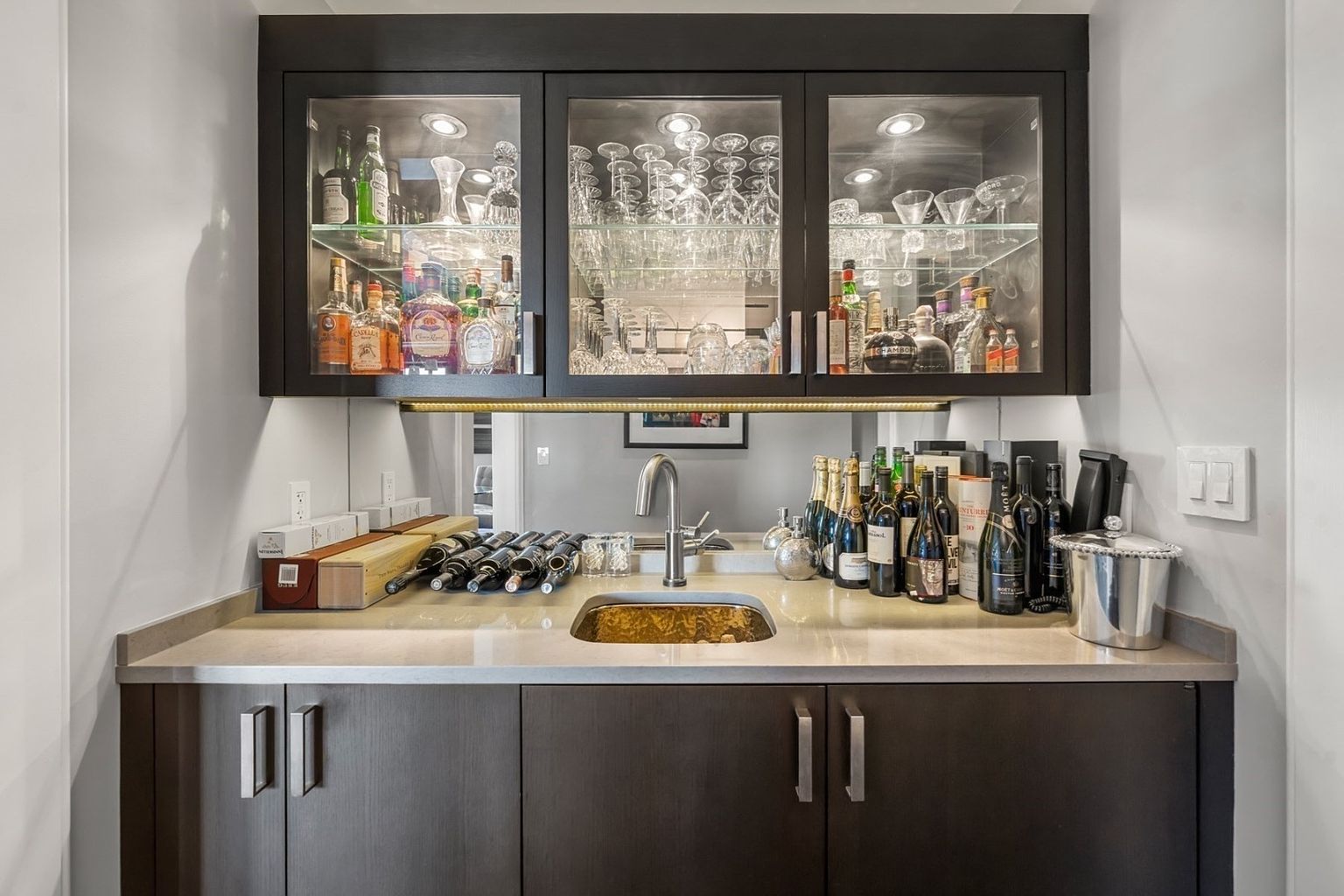 This image showcases a sophisticated wine cellar or bar area, featuring dark wood cabinetry with glass-fronted upper cabinets displaying an array of glassware and liquor bottles. Below, a countertop with a sink is lined with wine bottles and various bar accessories, illuminated by subtle lighting. The overall impression is one of luxury and refined taste, perfect for entertaining.