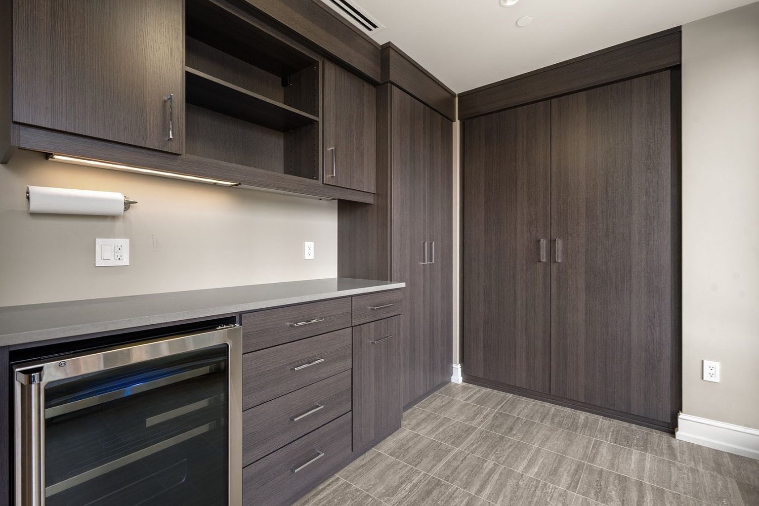 This interior shot showcases a modern wine cellar with dark wood cabinetry and a built-in wine refrigerator. The gray countertops and tiled flooring complement the sleek design. The space is well-lit, highlighting the storage capacity and sophisticated aesthetic, making it an attractive feature for wine enthusiasts.