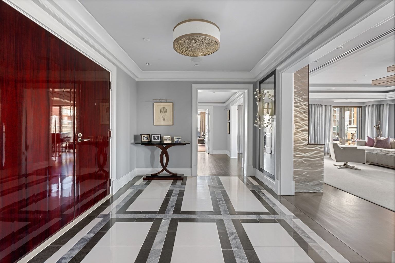This is an interior shot of a luxurious hallway featuring a striking geometric tile floor pattern in black, white, and gray. A high-gloss, dark wood door stands to the left, while a console table with framed photos sits against a light gray wall. The hallway leads to other rooms, offering a glimpse into a well-appointed living space, and a modern chandelier provides elegant lighting.