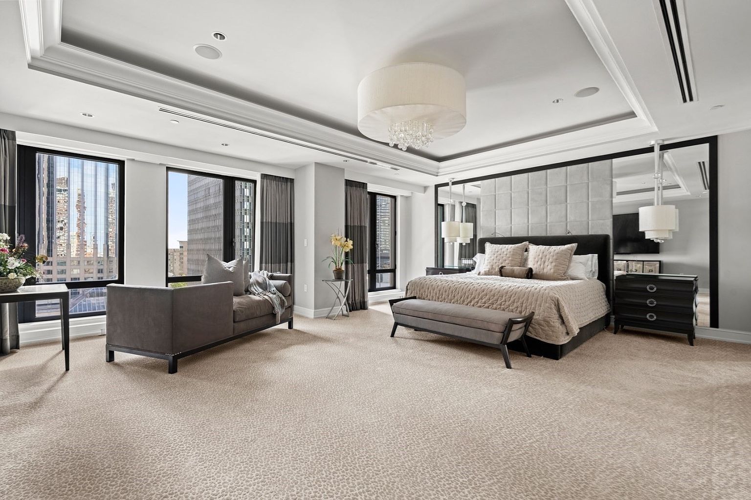 This is a spacious primary bedroom featuring a large bed with a tufted headboard and neutral-toned bedding. The room has large windows offering city views, a seating area with a gray sofa, and a modern chandelier. The overall style is luxurious and contemporary, with a focus on comfort and elegance.
