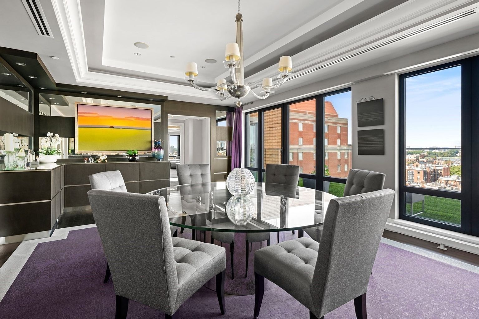 This is a dining room featuring a round glass table surrounded by six gray upholstered chairs. A modern chandelier hangs above the table, and large windows offer a city view. The room is decorated with a purple rug and artwork, creating a sophisticated and elegant atmosphere.