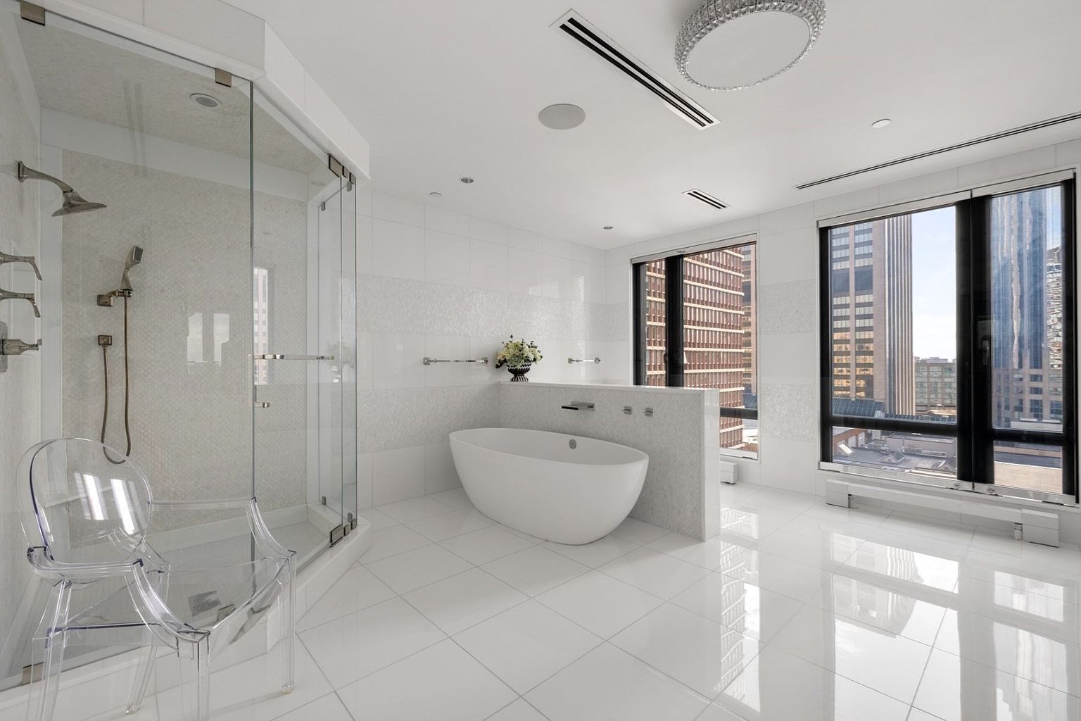 This is a luxurious primary bathroom featuring a modern freestanding bathtub, a glass-enclosed shower, and large windows offering city views. The bathroom is predominantly white with tile flooring and walls, creating a bright and clean aesthetic. A clear acrylic chair sits near the shower, adding a contemporary touch.
