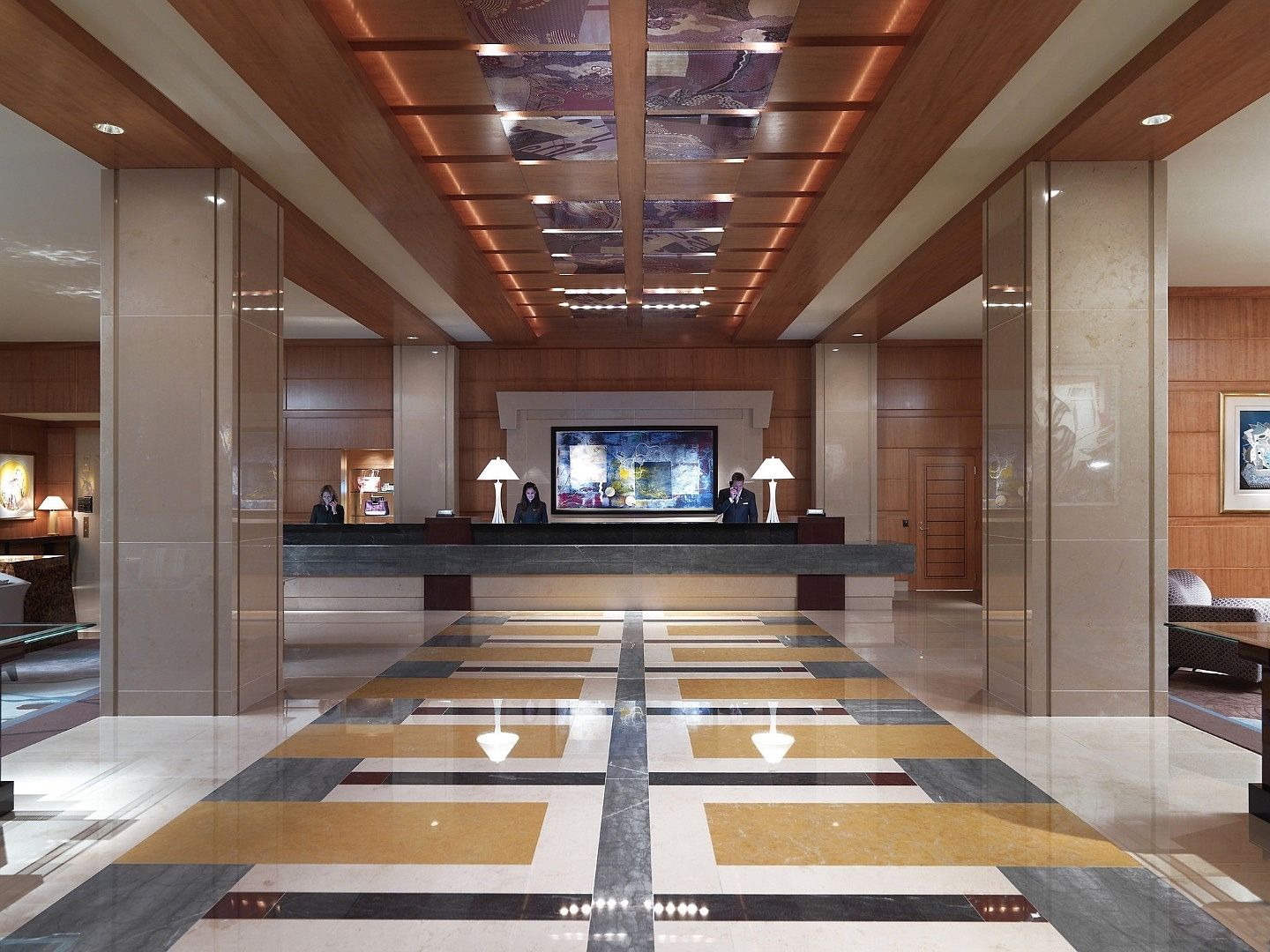 This is a grand hotel lobby featuring a long reception desk with staff, a patterned marble floor, and wood-paneled walls and ceiling. The lighting is warm and inviting, creating a luxurious and sophisticated atmosphere. The perspective is a straight-on view, emphasizing the length and symmetry of the space.