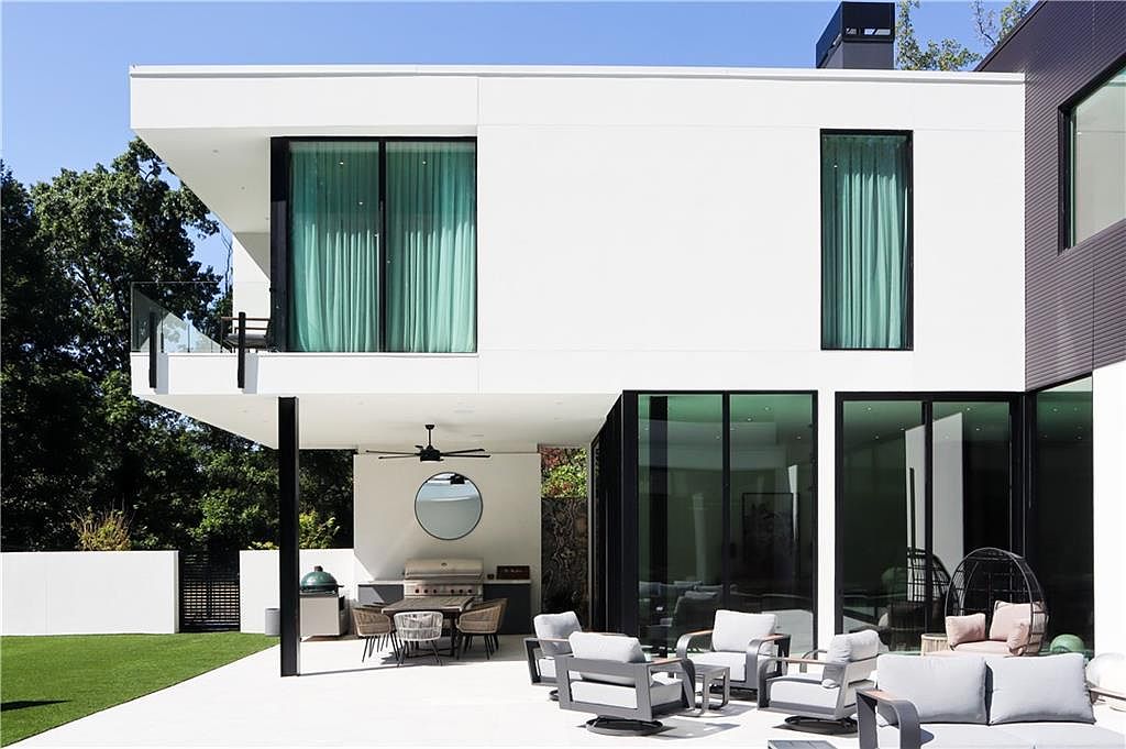 This modern rear exterior view showcases a sleek, white-stucco home with clean lines and expansive glass elements. The property features a spacious covered patio area equipped with an outdoor kitchen and dining set, transitioning into a comfortable lounge space with contemporary outdoor furniture. The design emphasizes indoor-outdoor living, highlighted by large sliding glass doors and a second-story balcony overlooking the manicured lawn.
