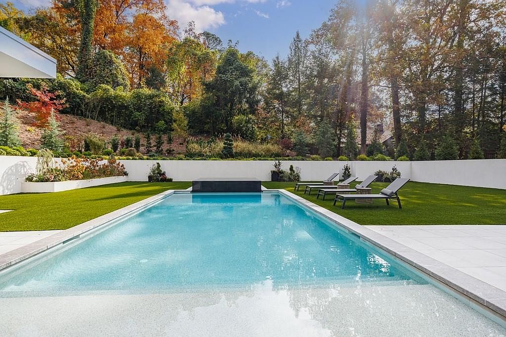 This image showcases a pristine, rectangular swimming pool set against a backdrop of lush, autumn-colored trees. The pool area features a clean, modern design with a light-colored stone deck, a dark water feature at the far end, and several lounge chairs arranged on a manicured artificial lawn. A white perimeter wall provides privacy, creating a serene and upscale outdoor retreat.