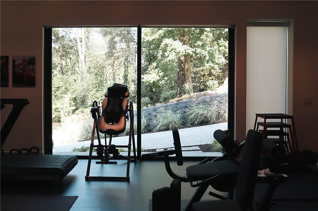 This home gym features a minimalist design with dark flooring and large, floor-to-ceiling windows that provide a clear view of the lush, wooded backyard. The space is equipped with various fitness machines, including an inversion table positioned centrally and a weight bench in the foreground. The natural light streaming through the expansive glass creates an airy, serene atmosphere for a workout.