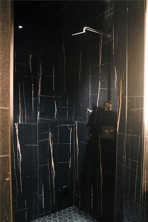 This modern shower enclosure features striking black stone wall tiles with prominent, light-colored veining, creating a dramatic and luxurious aesthetic. The space is equipped with a sleek, wall-mounted rainfall showerhead and a separate handheld sprayer, complemented by a minimalist control valve. The floor is finished with small, dark hexagonal tiles, providing a textured contrast to the smooth, large-format wall panels.